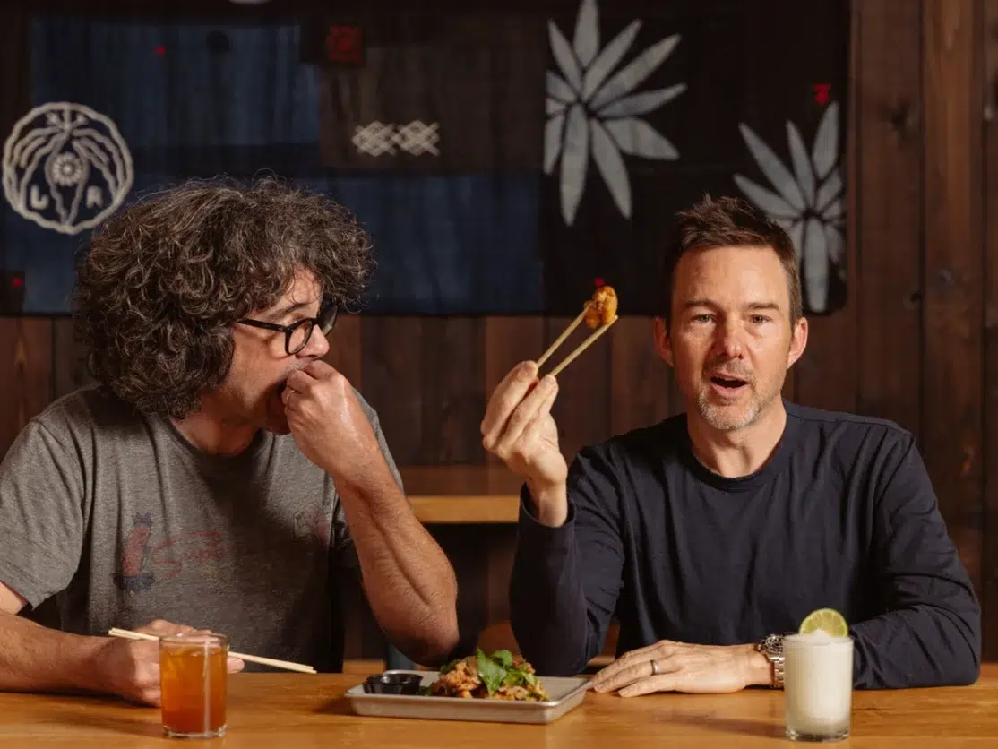 Aaron Franklin and Tyson Cole sitting at a table eating food with chopsticks.