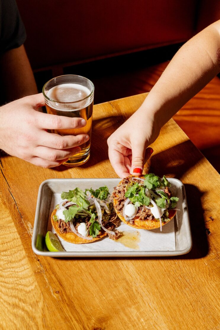 A person reaching for a brisket tostada on a white plate on a wooden table.