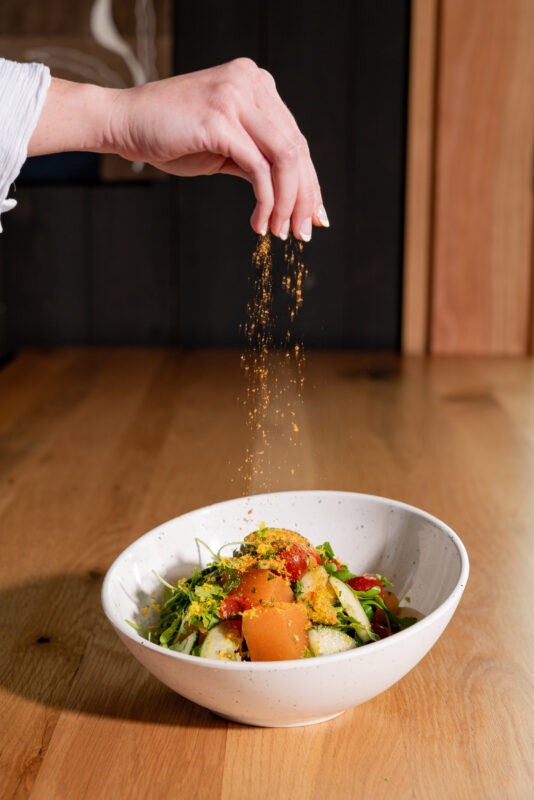 A chef sprinkling seasoning into a white bowl filled with a salad on top of a wooden table.