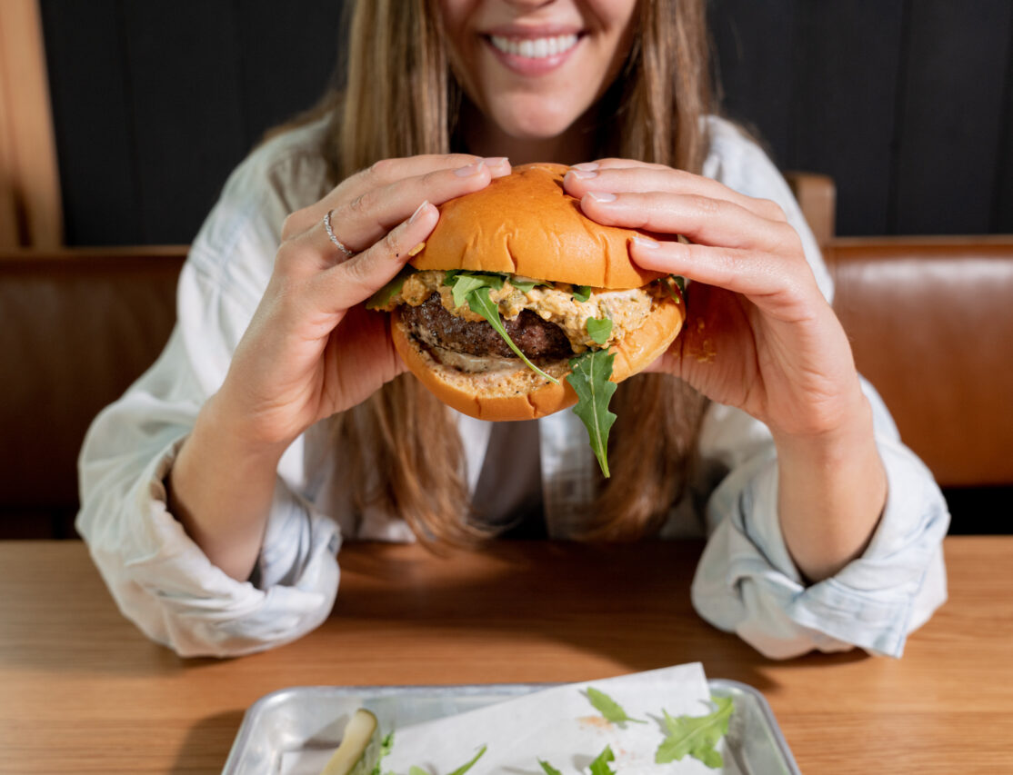 A woman sitting at a table with a caramelized onion-cheddar burger in front of her.