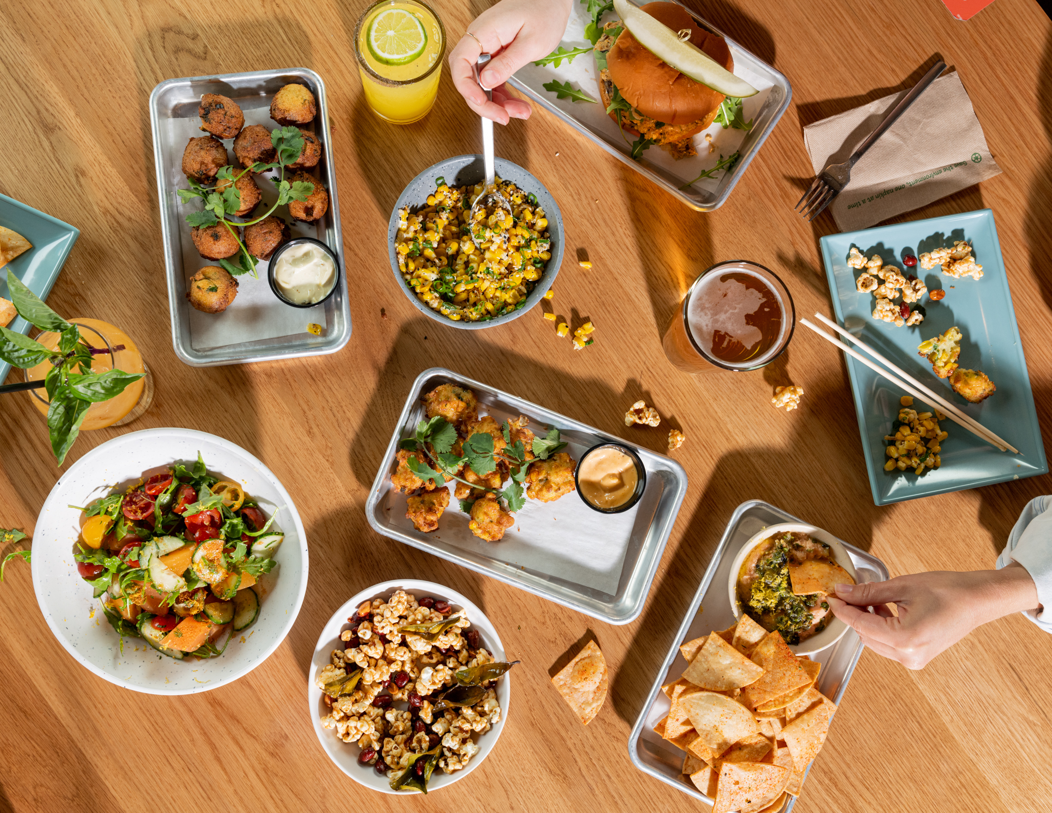 A wooden table topped with many plates of food, two cocktails, and a pint of beer.