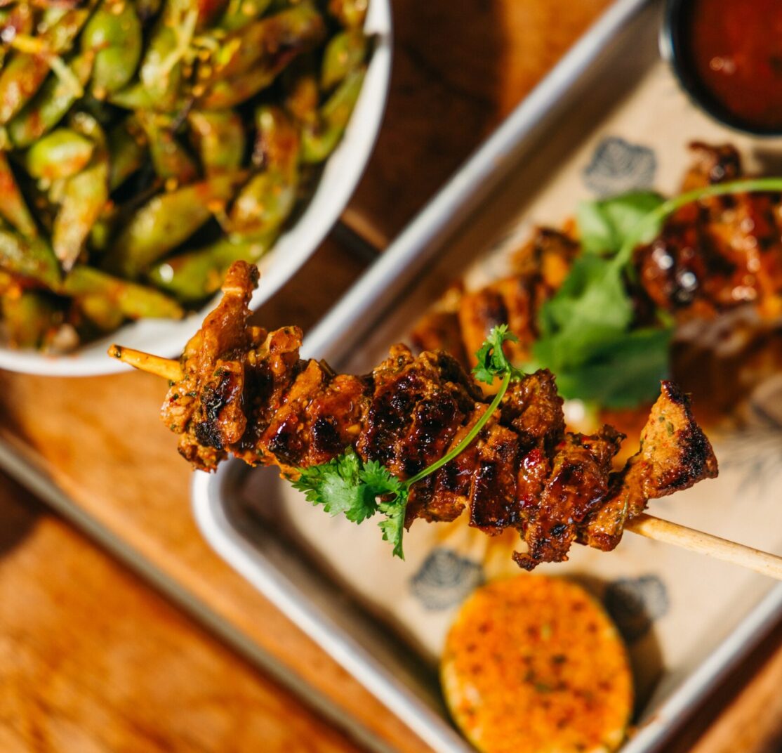 close up of hand holding charred pork skewer with happy hour spread behind
