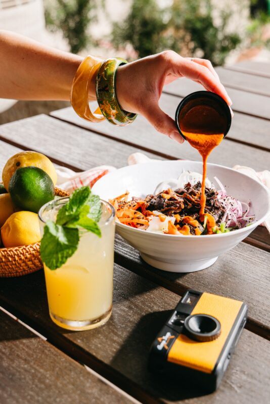 hand pouring orange Korean scallion sauce over charred mushroom bowl with yellow seasonal cocktail on the table