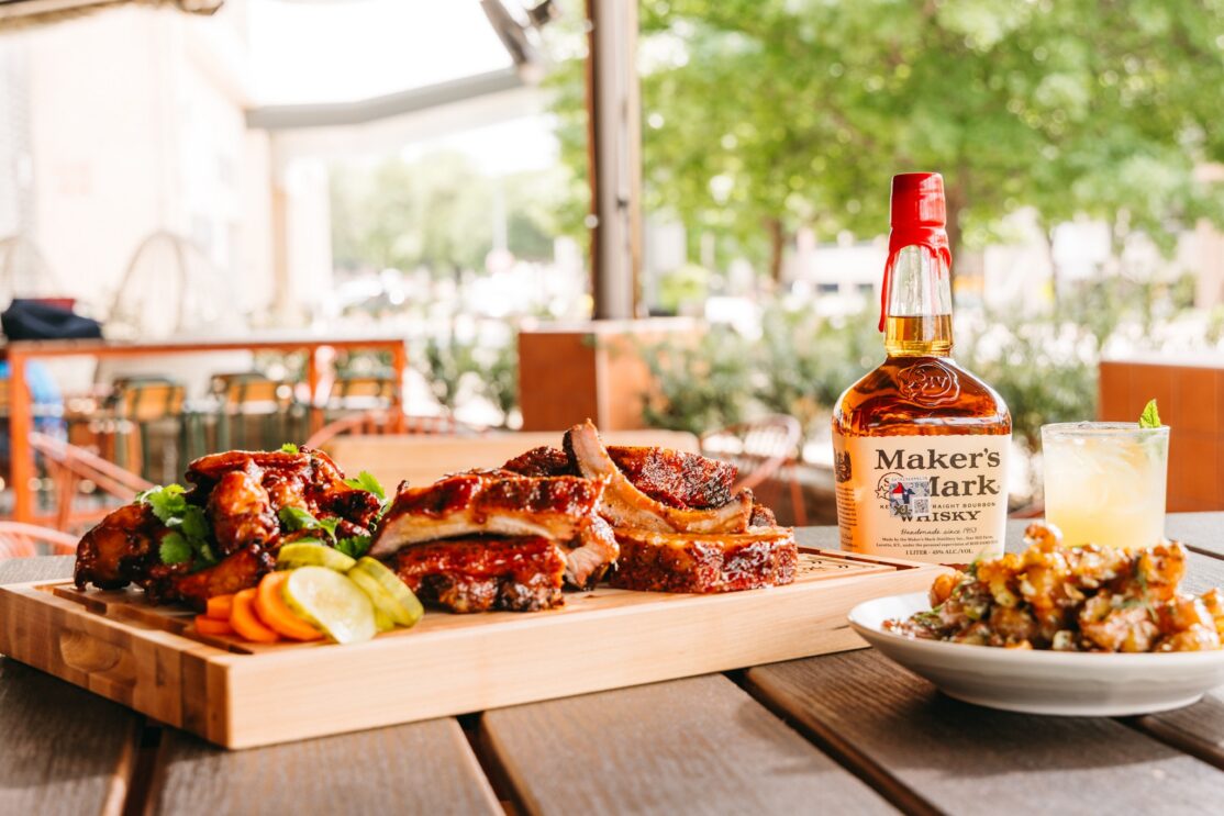 wooden platter of ribs and wings with maker's mark whiskey bottle next to a yellow bourbon cocktail